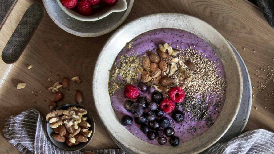 Brett mit Schüssel mit violettem Joghurt, getoppt mit Blaubeeren, Himbeeren, Mandeln und Saaten, daneben kleine Schälchen mit mehr Toppings