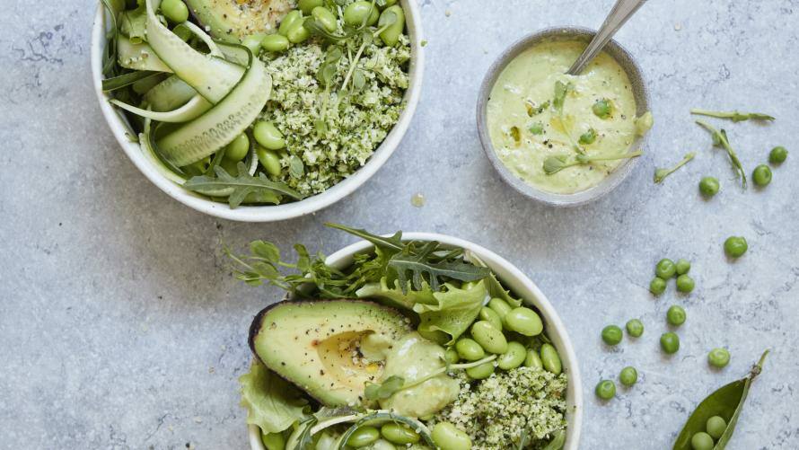 Zwei Schüsseln mit Brokkolireis, Gurkenstreifen, Avocado, Edamame und verschiedenen Blattsalaten, daneben Schälchen mit Erbsendressing