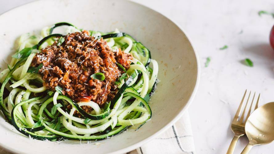 Zucchini-Nudeln mit Bolognese in einem großen Teller mit Besteck und Tomaten