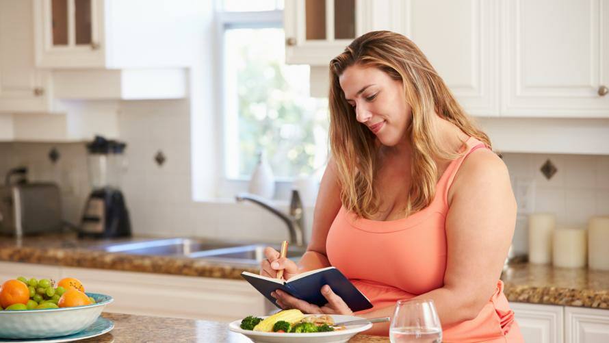 Frau schreibt in der Küche sitzend in ihr Ernährungstagebuch