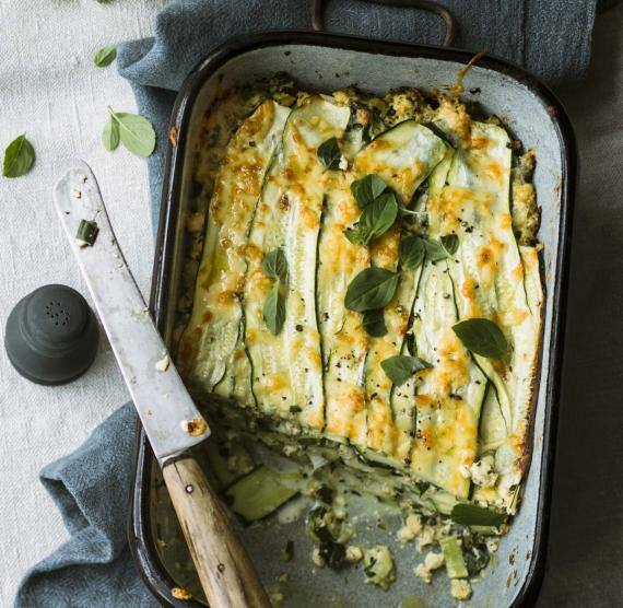 Auflaufform auf Tuch mit angeschnittener Zucchini-Lasagne mit Käse überbacken und Basilikumblättern bestreut, daneben schwarzer Salzstreuer und Messer