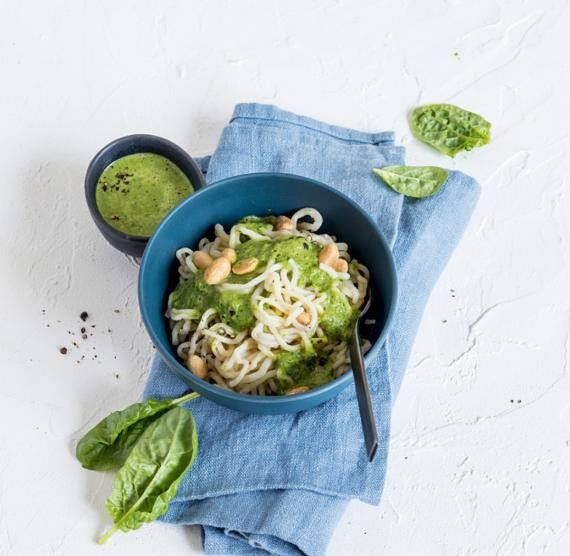Dunkle Schüssel mit Konjaknudeln, grünem Pesto und Sojabohnenkernen, daneben ein Schälchen mit mehr Pesto und einzelnen Spinatblättern