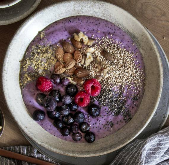 Brett mit Schüssel mit violettem Joghurt, getoppt mit Blaubeeren, Himbeeren, Mandeln und Saaten, daneben kleine Schälchen mit mehr Toppings
