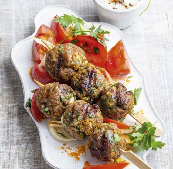 Tablett mit zwei Hackbällchen-Spießen, daneben Tomatensalat und ein Schälchen mit Joghurt-Dip, außerdem: Petersilienzweige