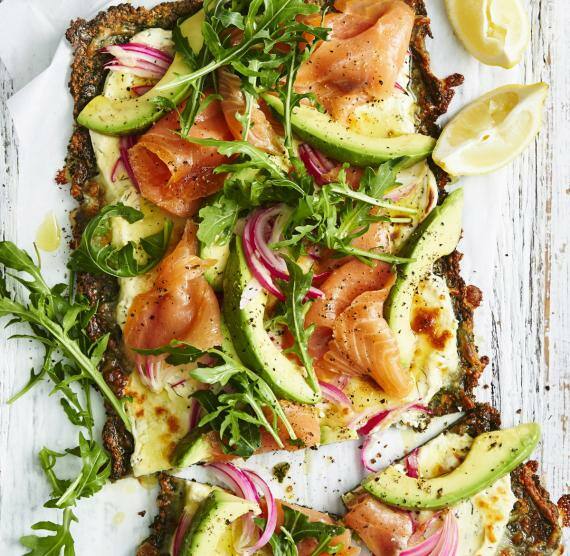 Pizza auf hellem Untergrund mit Frischkäse, roten Zwiebeln, Räucherlachs, Avocado und Rucola, darüber schwarzer gemahlener Pfeffer