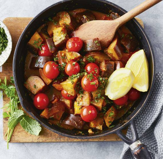 Beschichtete Pfanne mit Auberginenstücken und Tomaten, darauf gehackte Kräuter und zwei Zitronenspalten, daneben mehr Kräuter