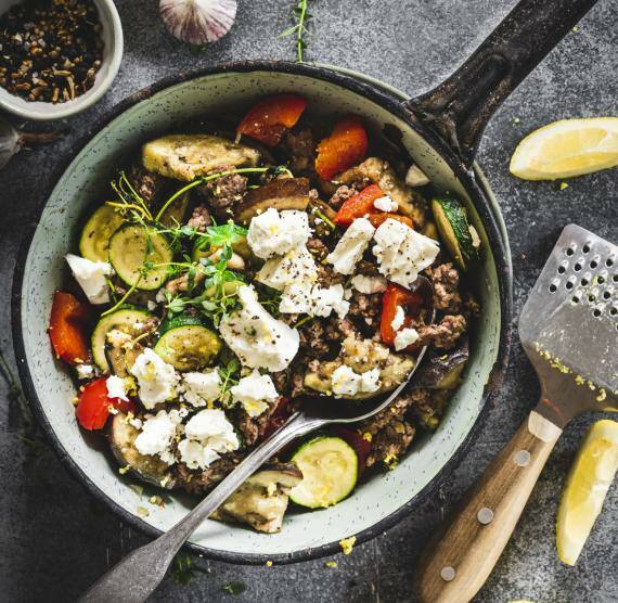 Pfanne mit Hackfleisch, Paprika, Feta, Zucchini, Aubergine und Zitronenspalten, darüber Kräuterzweige
