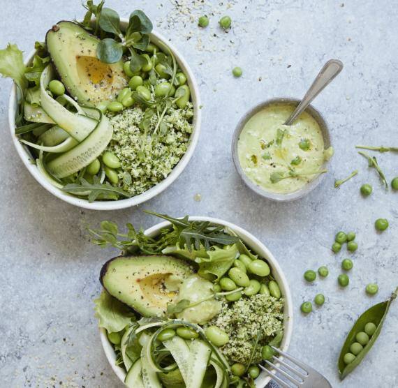 Zwei Schüsseln mit Brokkolireis, Gurkenstreifen, Avocado, Edamame und verschiedenen Blattsalaten, daneben Schälchen mit Erbsendressing