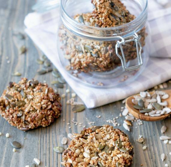 Saaten-Cracker auf einem Tisch und im Glas, außerdem einzelne Sonnenblumen- und Kürbiskerne sowie Sesam und gehackte Mandeln