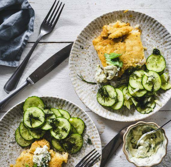 Backfisch mit Gurkensalat auf zwei Tellern mit Besteck, daneben Remoulade