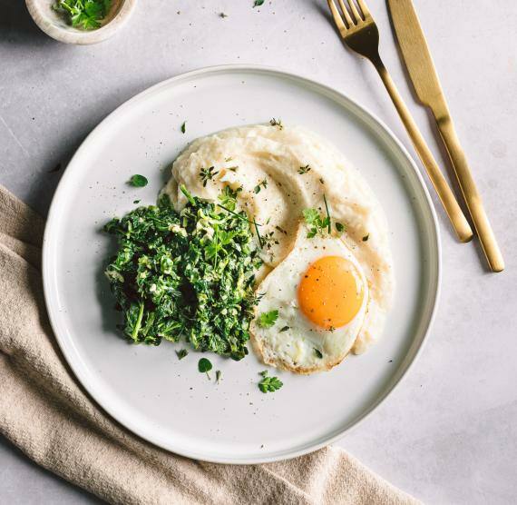 Blumenkohlpüree mit Rahmspinat und Spiegelei auf einem Teller mit goldenem Besteck
