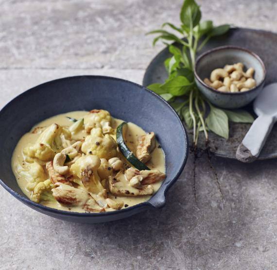 Curry in einem tiefen, dunklen Teller, daneben Kräuter und Cashews