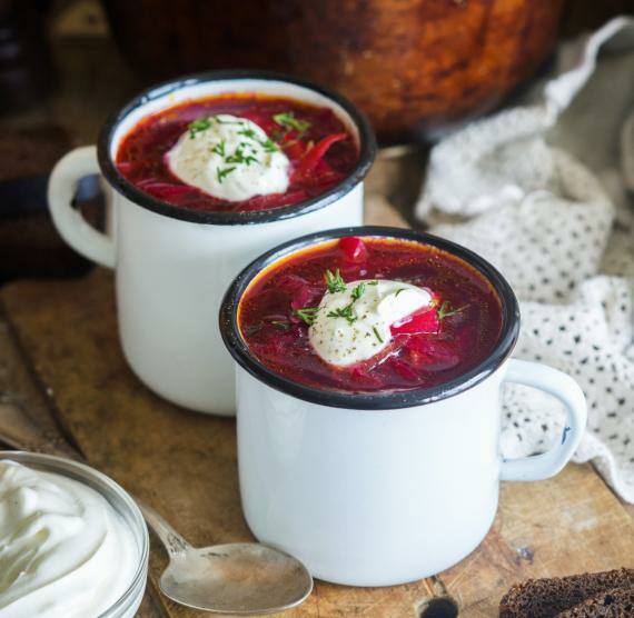 Rote Bete-Suppe in zwei Emaille-Tassen, dahinter ein großer Topf