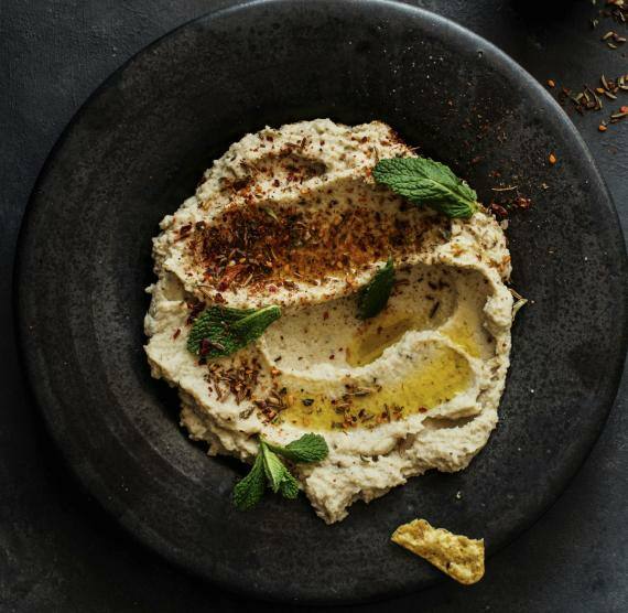 Schwarzer Teller mit beige-farbenem Dip, darüber rote Gewürze, Öl und Minze