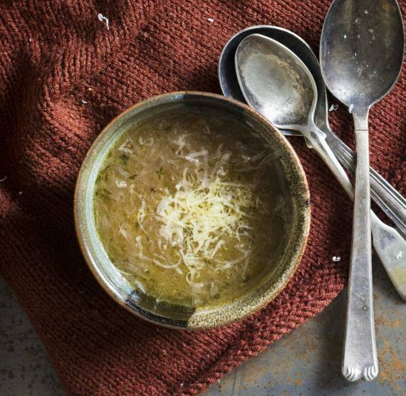 Schälchen mit Suppe auf rostrotem Strick, daneben ein Stück Käse mit einer Reibe