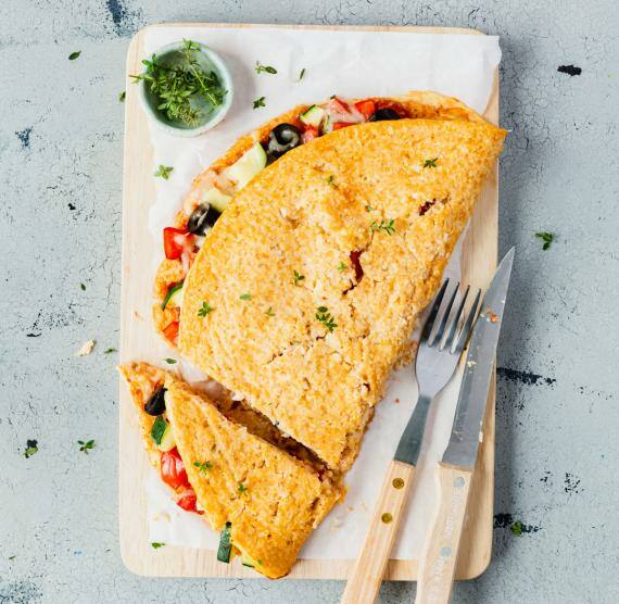 Brett mit Low Carb Blumenkohl-Calzone, gefüllt mit buntem Gemüse, daneben Kräuter