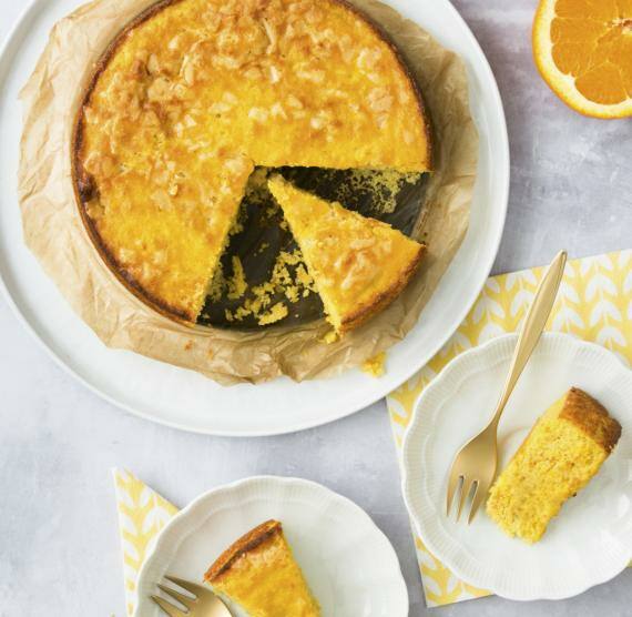 Mallorquinischer Mandelkuchen auf einer Kuchenplatte, daneben zwei Stücke auf Tellern und eine halbierte Orange