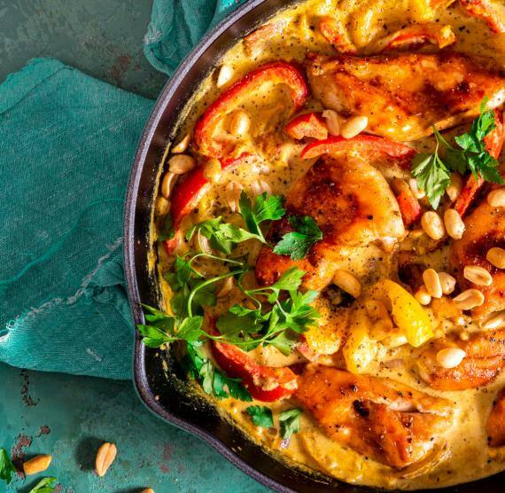 Pfanne mit Hähnchen, Paprika und Erdnusssauce auf einem Tisch, daneben Erdnüsse